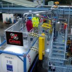 Two men stand on a Carbis Solutions elevated truck platform above a tank truck.