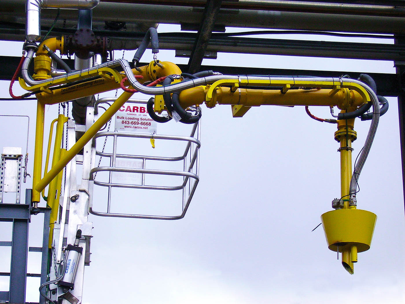 A loading arm is coming down from a pipe rack, positioned in front of a telescoping safety cage.