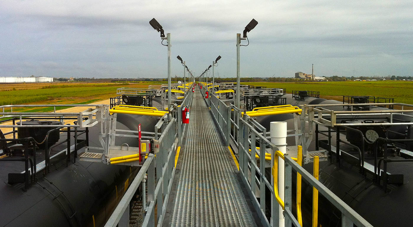 Gangways fall out on both sides of a catwalk resting above 2 railroad tracks. Railcars are accessible and underneath them.