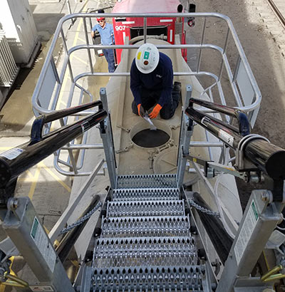 A man works on the top of tanker truck in a gangway cage below folding stairs.