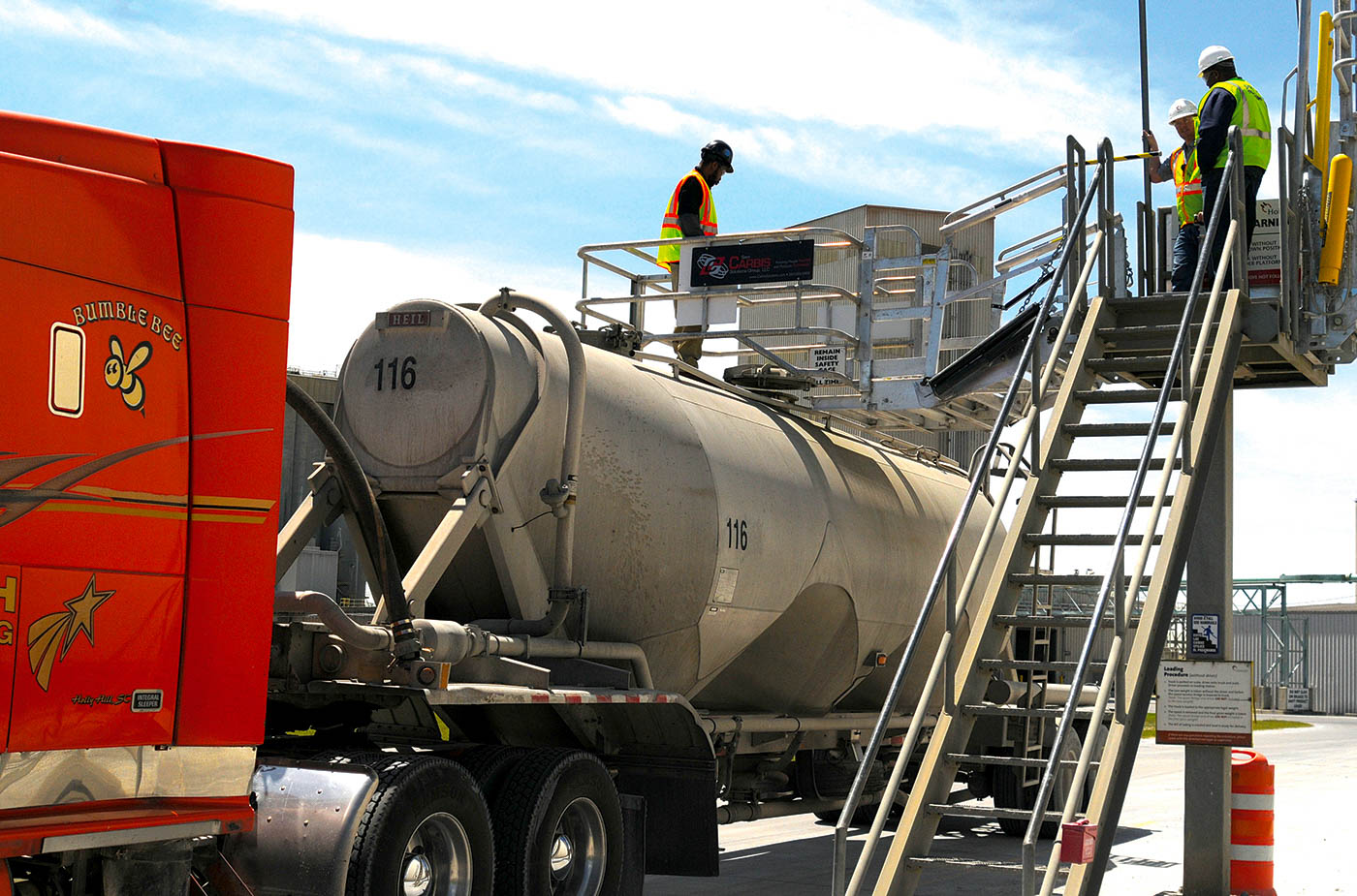 A truck safety cage sits atop a tanker truck. A worker is positioned on it.